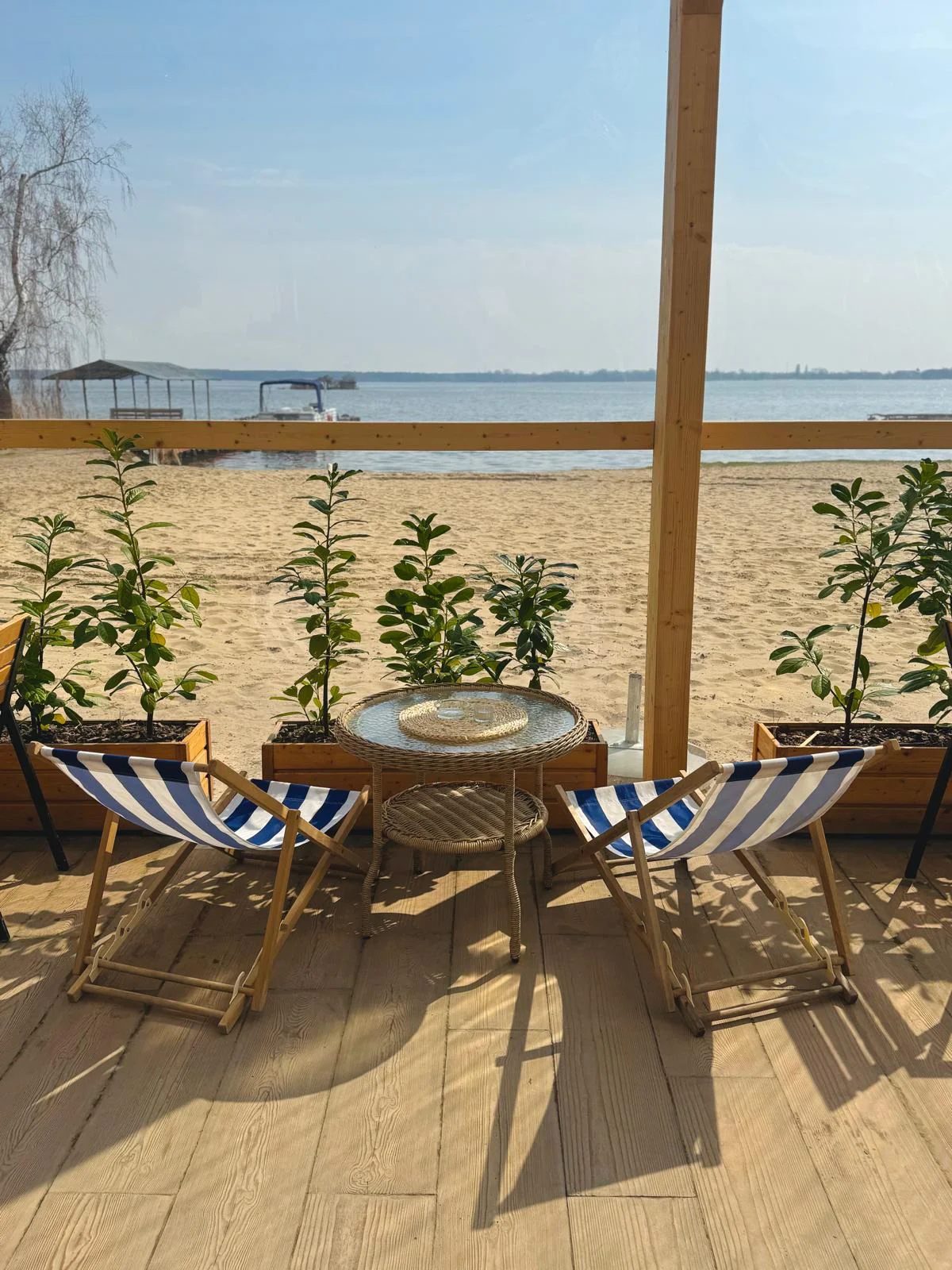 Dwa leżaki w granatowo-białe paski na drewnianym tarasie, za nimi plaża i jezioro