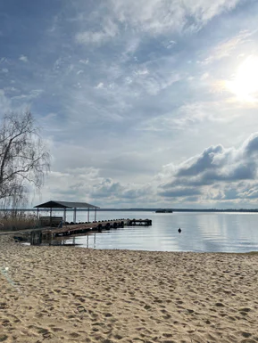 A wide sandy beach and pier on the lake, with sun and clouds above