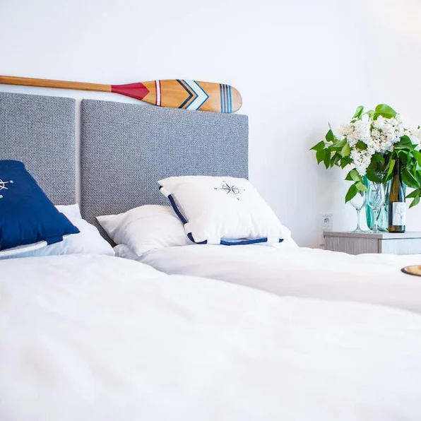 Bed with white linen and nautical cushions, an oar above it and a bunch of lilac beside it